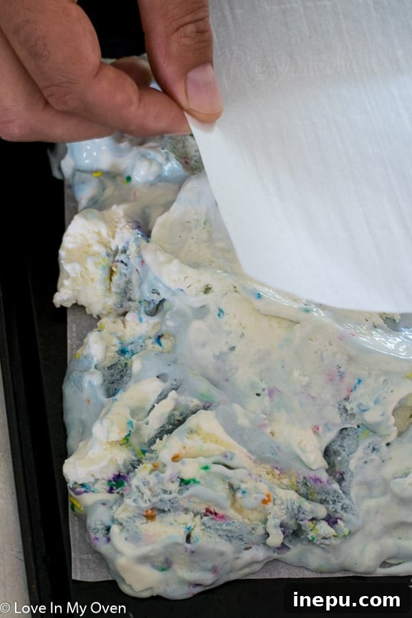 A close-up of a perfectly smooth layer of birthday cake ice cream between two sheets of parchment paper, ready for freezing.