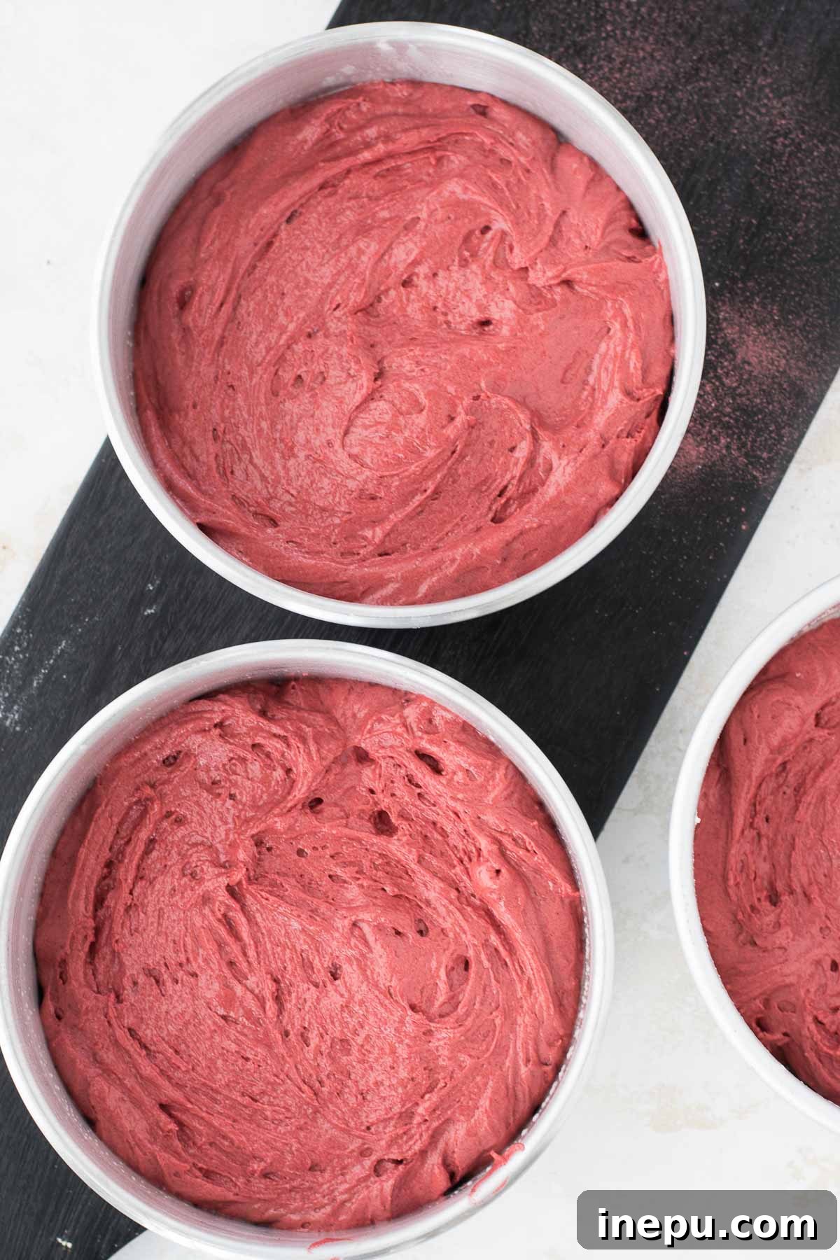 Three cake pans filled with red velvet batter, ready for baking.