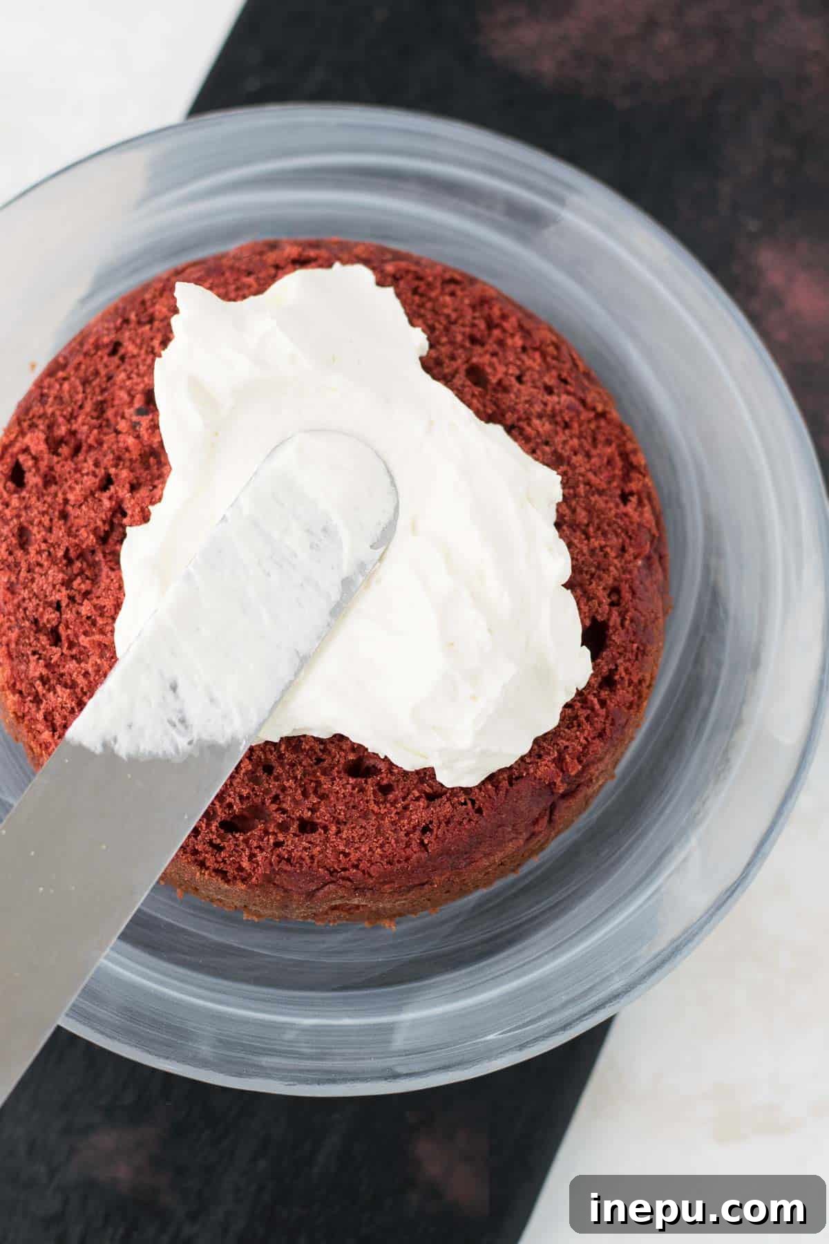 A mini red velvet cake being frosted with white whipped cream cheese frosting on a rotating cake stand.
